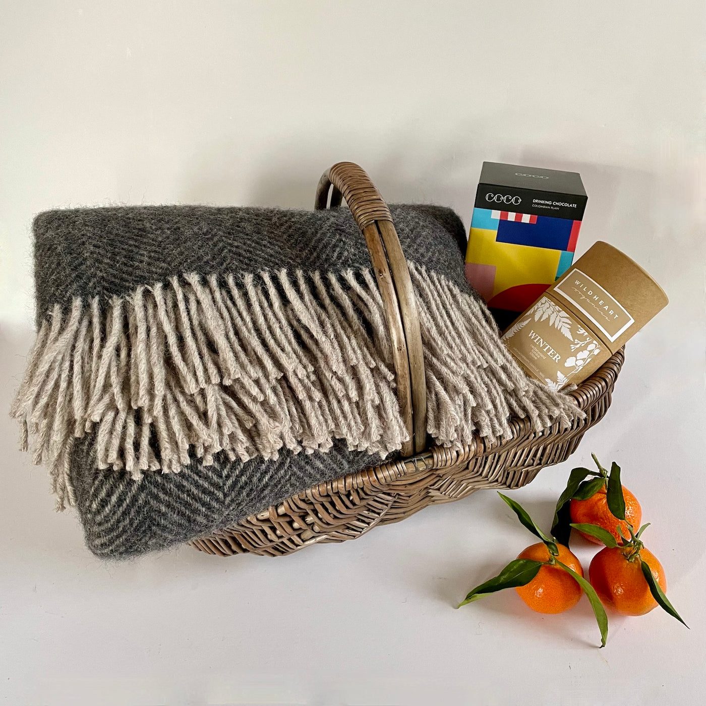 The St Andrews Hamper Company hygge pamper hamper with a selection of organic and eco-friendly gifts including a charcoal and silver herringbone weave pure new wool full size throw, Wildheart Organics winter scented natural soy candle, Coco Chocolatier Columbian drinking chocolate inside a hessian lined handwoven country style basket.